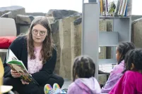 Une personne adulte assise par terre lit un livre à trois enfants assis devant elle.