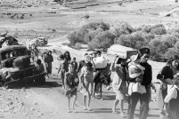 Une photo en noir et blanc d’un groupe d’adultes et d’enfants transportant leurs affaires, marchant sur une route désertique non pavée près d’un camion en ruine.