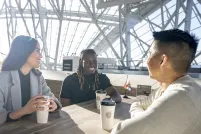 Trois personnes sont assises à une table. Elles sourient toutes, un café devant elles. Un petit drapeau de la Fierté progressif est posé sur leur table et sur la table vide derrière elles. Le soleil brille à travers les fenêtres du Musée.