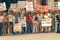 Un grand groupe de personnes vêtues de t-shirts et de pantalons évasés à la mode des années 1970 sont debout ensemble et tiennent des pancartes portant des messages en anglais tels que « sortis du placard », « c’est bien d’être gai » et « abrogez les lois anti-gaies ».