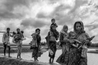 Un groupe de femmes et d’hommes rohingyas transportent de jeunes enfants et des effets personnels en marchant sur une digue de terre qui traverse une étendue d’eau.