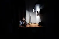 Un homme blanc aux cheveux courts est assis seul à un bureau illuminé dans une pièce sombre. Il fixe un écran d’ordinateur.