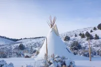 Un tipi recouvert de neige se dresse dans un paysage vallonné parsemé d’arbres et de buissons, sous un ciel bleu.