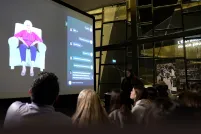 Un auditoire regarde un grand écran de projection sur lequel apparaît une femme âgée vêtue d’un haut violet et d’un pantalon bleu, assise dans un fauteuil blanc, à côté d’une interface interactive. La présentation se déroule dans un espace moderne à l’architecture géométrique en verre, et une personne se tient à droite de l’écran.