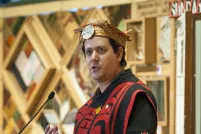 Un homme vêtu d’un costume et d’une coiffe traditionnels autochtones se tient devant une installation d’art faite de panneaux de bois avec des objets encastrés.