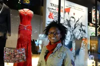Une femme souriante se tient devant une robe de bal rouge et un smoking noir exposés sur des mannequins. La robe et le costume sont derrière une vitrine.