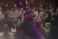 Un groupe d’hommes placés en cercle joue du tambour et chante. Une foule nombreuse et diversifiée les entoure de près et les accompagne en chantant.