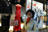 Une femme souriante se tient devant une robe de bal rouge et un smoking noir exposés sur des mannequins. La robe et le costume sont derrière une vitrine.