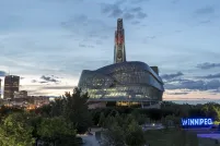 Photo extérieure des fenêtres en verre et de la tour du Musée canadien pour les droits de la personne, prise au crépuscule avec le ciel des prairies en arrière-plan. Le panneau Winnipeg à Forks est situé à droite de l'image. Plusieurs tours de bureaux et les lumières du centre-ville sont visibles à gauche de l'image.