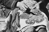 Une photo en noir et blanc montrant des mains tenant et cousant des morceaux de tissu décorés de broderies florales et de fibres texturées, étalés sur les genoux d’une personne.