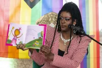Une drag queen très maquillée et coiffée d’une perruque composée de longues et minuscules tresses brandit un livre pour enfants pour le montrer à un public. La toile de fond derrière elle est un arc-en-ciel pastel.