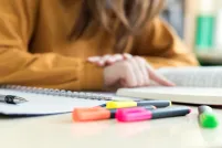 Gros plan sur un cahier à spirales, un stylo et des surligneurs colorés posés sur un bureau, avec, à l’arrière-plan, une personne vêtue d’un chandail couleur moutarde, dont la silhouette est floue.