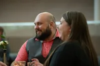 Un homme et une femme sont assis ensemble à une table, souriant chaleureusement tout en écoutant d’autres convives. L’homme porte une chemise saumon et un gilet gris, et de petites bougies et des boissons sont disposées sur la table, créant une atmosphère intime et chaleureuse.