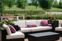 Des canapés et des chaises sont installés dans un patio clôturé. Le patio est entouré de petits arbres et de hautes herbes vertes de la prairie. En arrière-plan, la flèche de l'Esplanade Riel s'élève au-dessus des arbres.