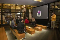 Un petit groupe de personnes assises sur des bancs en bois dans un théâtre fait face à un écran montrant une femme âgée assise dans un fauteuil. Un homme vêtu d’une chemise jaune se tient à droite de l’écran et tient un microphone.