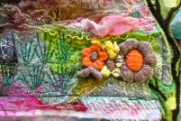 Une œuvre d’art textile colorée qui comprend des fleurs brodées, des branches et de la verdure mélangées à des couches de tissus roses, bleus et blancs. 