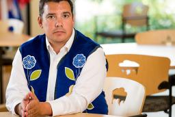 A man wearing a blue vest with flower beadwork, seated with his hands resting on a table.