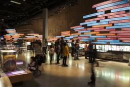 Un grand groupe de personnes dans une galerie dont un mur est recouvert de panneaux colorés et de photos montrant une chronologie. 