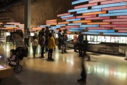 Un grand groupe de personnes dans une galerie dont un mur est recouvert de panneaux colorés et de photos montrant une chronologie. 