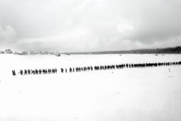 Une photo en noir et blanc d’un paysage plat et enneigé. On voit des arbres et des petits bâtiments au loin. Une longue file de plus de 100 personnes marchent dans la neige.