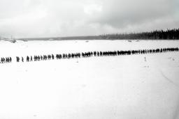 Une photo en noir et blanc d’un paysage plat et enneigé. On voit des arbres et des petits bâtiments au loin. Une longue file de plus de 100 personnes marchent dans la neige.