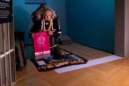 A man wearing an eagle-feather headdress, holding a pipe, kneels over a fur pelt in a room that resembles a small prison cell.