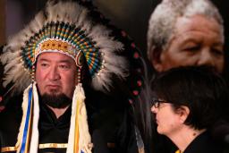 A man wearing a feather headdress stands beside a woman, who is looking at him. There is a large photograph of a man with grey hair (Nelson Mandela) in the background.