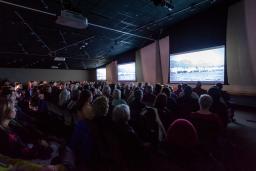 Un auditoire assis regarde trois grands écrans dans une salle dont l'éclairage est tamisé.