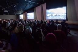 Un auditoire assis regarde trois grands écrans dans une salle dont l'éclairage est tamisé.