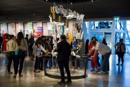 Un grand groupe de personne dans une galerie examine un élément d’exposition comprenant plusieurs objets d’apparence ordinaire.