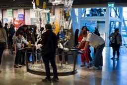 Un grand groupe de personne dans une galerie examine un élément d’exposition comprenant plusieurs objets d’apparence ordinaire.