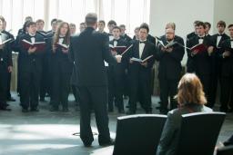 Une chorale d’hommes habillés de noir, tenant des livres et chantant devant un chef de chœur.