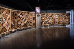 Une grande installation artistique horizontale à armature en bois composé de multiples losanges et rectangles sur lesquels sont fixés de nombreux objets. Une porte est entrouverte en plein centre de l’œuvre.