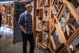 Two people look at a large artwork consisting of objects set in cedar frames, with an open door in the middle.