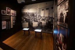 Une exposition muséale montrant une photo en noir et blanc d’enfants assis à des pupitres placés en rangées. Deux pupitres semblables à ceux qu’on voit dans la photo sont placés au centre de l’exposition. Sur un panneau de texte, on peut lire « Enfances oubliées ».