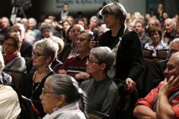 Une salle remplie de personnes assises. Une femme est debout et tient un microphone portable.