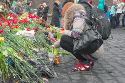 Deux personnes déposant des fleurs rouges à un mémorial. En arrière-plan, une douzaine d’autres personnes se tiennent devant des débris, comme des roches, des feuilles de métal et des barrières de ciment peintes avec des drapeaux ukrainiens bleu et jaune et des lettres.