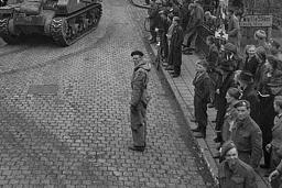 Une foule assemblée sur des trottoirs et regardant des véhicules blindés rouler sur les rues pavées d’un village.
