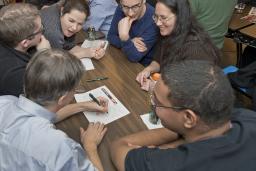 Des gens enthousiasmés regroupés autour d’une table. Un homme écrit sur une feuille de papier.