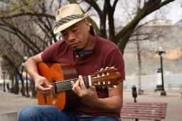 A man wearing a fedora plays a guitar while sitting outside on a bench. There are trees in the background.