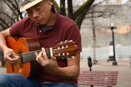 A man wearing a fedora plays a guitar while sitting outside on a bench. There are trees in the background.