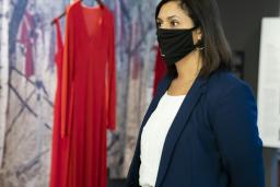 A woman wearing a black face mask stands in front of a museum exhibit consisting of red dresses hanging in front of a woodland background.