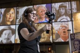 A woman wearing headphones, holding a mobile device on a handle, stands in a Museum gallery.