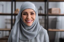 A woman wearing a blue hijab smiles toward the camera, with a bookshelf in the background.