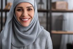 A woman wearing a blue hijab smiles toward the camera, with a bookshelf in the background.