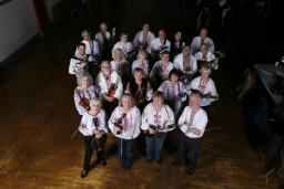A group of twenty-two people facing the camera and smiling. They are all wearing dark-coloured pants and white shirts with red embroidery. A majority of the group are holding their instruments, mainly mandolins. 