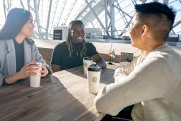 Three people sit around a table, smiling, each with a paper coffee cup in front of them. The sun shines behind them, through the panes of glass that wrap around the Canadian Museum for Human Rights.