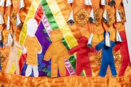 A section of an orange jingle dress is shown. Jingles, or small silver cone-shaped pendants, are visible at the top of the image. Under them, seven human figures are shown, all in various colours and sizes. One of the figures is wearing an orange shirt. These human figures are holding hands inside a multicoloured tipi.