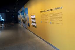 A yellow wall with the words "Ukrainian Artists United" and a dynamic blue and yellow graphic is right side of the image. On the left side, an image is projected on a screen showing a person with headphones and an open mouth, as though they are singing. Benches for sitting are placed in front of the screen.