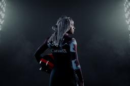 A black woman stands in a dark place between two large projectors. She's holding a helmet and wears a Canadian bobsleigh suit.